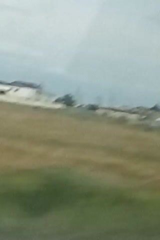 A grainy, low-resolution video captures a view from a moving vehicle. The scene appears to be a rural or semi-rural area with dry grass in the foreground. In the background, there are several buildings, some with blue roofs and others with lighter colored walls. Some structures appear to be industrial or utility buildings, with tall poles or stacks visible. The sky is overcast. The video quality is poor, making it difficult to discern specific details about the buildings or any potential occupants.