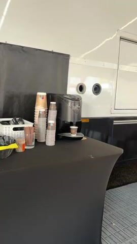 The video shows a coffee machine dispensing coffee into a small paper cup. A stack of paper cups is visible to the left of the coffee machine, along with a white basket containing utensils. The scene appears to be outdoors, possibly at an event, with a black tablecloth covering a table. The background shows part of a white trailer.