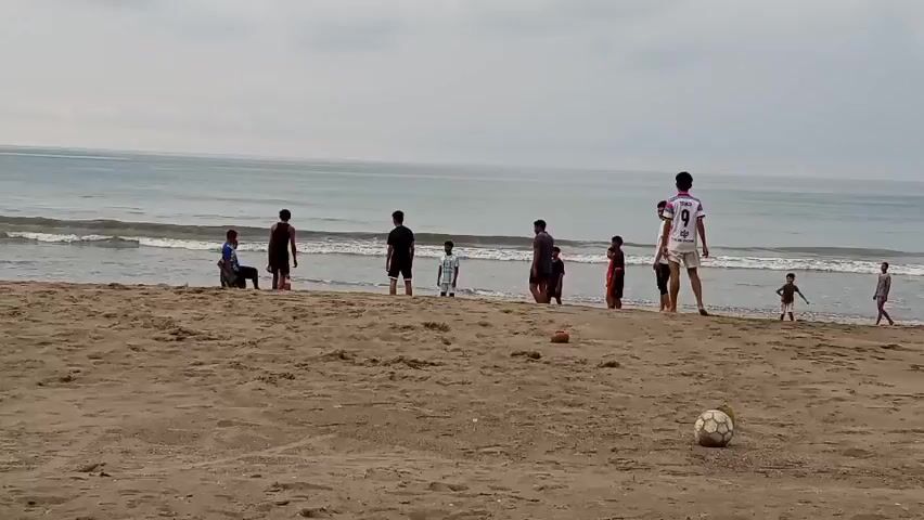 A group of boys and young men are playing soccer on a sandy beach near the ocean. The sky is overcast, and the waves are gentle. Several boys are in the water, while others are on the sand, kicking and chasing a soccer ball. One boy in a white and pink jersey appears to be kicking the ball high in the air, while others are positioned around the beach, ready to receive it or play defense. There are at least ten individuals visible in the video, and two soccer balls are on the sand. The scene is casual and appears to be a friendly game of soccer on a warm day.
