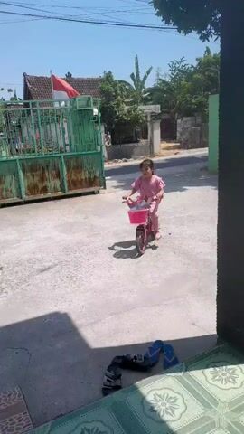 A young girl in a pink shirt and pink pants rides a pink bicycle with a pink basket. She is on a paved surface, possibly a street or driveway. In the background, there is a green fence and some trees. There is also a flag flying, which appears to be red and white. A motorcycle is visible in the background as well, with a rider. Two pairs of flip-flops are on the ground in the foreground.