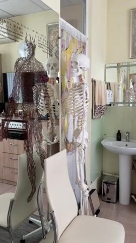 A video opens on a room with a mirror and a skeleton. Two skeletons are visible, one in the foreground and another reflected in the mirror. The skeleton in the foreground is a full anatomical model. A colorful curtain hangs behind the foreground skeleton, displaying an abstract pattern of hands. The room appears to be a medical office or a similar professional space, with a sink and a counter visible in the background, along with medical charts or posters on the wall.