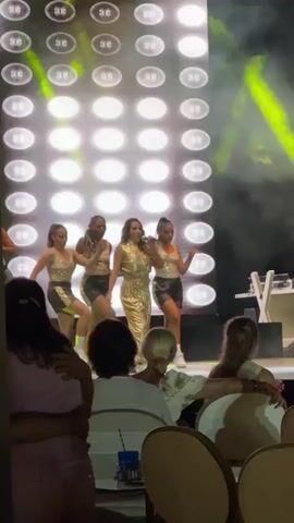 The video shows a live performance taking place on a stage in front of an audience. The main subject is a woman in a sparkly gold outfit, singing into a microphone and dancing. She is surrounded by several backup dancers also in gold and black outfits, performing synchronized movements. Behind the performers is a large screen or backdrop adorned with a grid of illuminated circles. Green spotlights are visible, adding to the stage lighting.

The audience members in the foreground are seated, facing the stage. Their backs are mostly to the camera, but their presence indicates a live event. The atmosphere appears energetic and festive, typical of a concert or show. The lighting suggests the event is taking place indoors or at night. There is no visible text in the video. The overall scene is one of entertainment and performance.