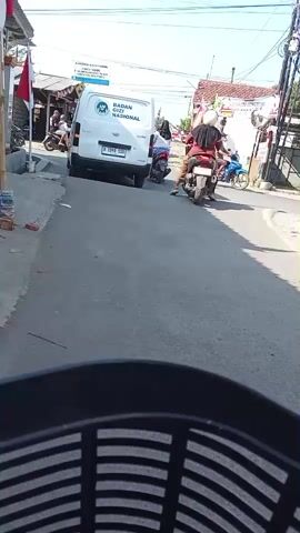A white van with the words BADAN POM NASIONAL printed on the back is parked on the side of a paved road. Two people on a scooter are stopped behind the van, and another person on a scooter is stopped in front of the van. A few other people are walking around on the sidewalk. The scene is outdoors, and the sun is shining.
