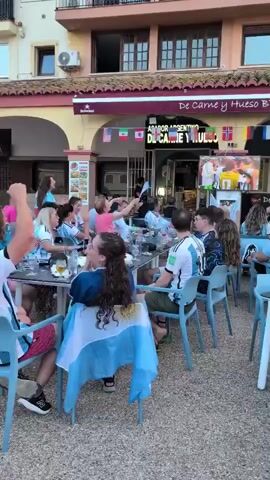 This media file captures an outdoor dining scene at a restaurant called "De Carne y Hueso BBQ" in Alboraia, Spain. A group of people, many wearing Argentina jerseys and flags, are gathered at tables, watching a televised event. The atmosphere appears festive and energetic, with some individuals raising their arms and appearing to cheer.

The subjects are primarily people seated at tables, engaged in dining and watching the screen. Objects include tables, chairs, drinks, and food items. The scene is set in an outdoor patio area of a restaurant, with a building providing a backdrop. The restaurant has an awning with flags from various countries displayed, including Argentina, Mexico, and Norway. Above the entrance, the restaurant's name, "De Carne y Hueso BBQ," and "ASADOR ARGENTINO," are visible.

The activity taking place is the observation of a sporting event or competition on a large screen, likely a football match given the Argentina national team colors and jerseys. The time of day appears to be late afternoon or early evening, as suggested by the lighting and shadows. The weather seems pleasant, conducive to outdoor seating.

Notable details include the prominent display of Argentina's national symbols, indicating support for the team. The presence of multiple national flags suggests an international context or a gathering of people from different backgrounds celebrating. The emotions conveyed are excitement, joy, and possibly anticipation. The text visible includes the restaurant's name and the type of cuisine offered.