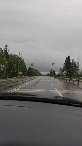 The video, shot from inside a car, shows a wet road during a rainy day. Raindrops are visible on the windshield, distorting the view of the overcast sky. The road is a two-lane highway, with trees lining either side. There are pedestrian crossing signs along the road, with white and yellow stripes marking the road surface. Streetlights are visible along the roadside. In the distance, the road continues to stretch, disappearing in the rain and clouds. The music is playing inside the car.