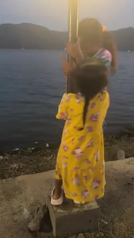 Two young girls are playing near a lamppost next to a body of water. The girl on the left, wearing a striped long-sleeved shirt and teal pants, is standing on a raised concrete block and holding onto the lamppost. She is smiling and looking at the other girl. The girl on the right, wearing a yellow dress with floral patterns, is also standing on the concrete block and holding onto the lamppost. She is looking up at the other girl and appears to be laughing. Both girls are wearing white clogs. The lamppost is dark and appears to be lit from above. The body of water is dark and calm, with hills visible in the background. The sky is dusky and overcast. There is some vegetation on the ground near the water's edge.