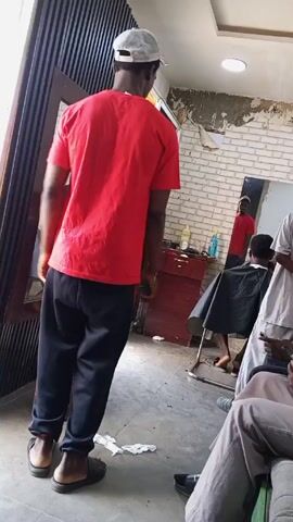 The video shows the interior of what appears to be a barbershop.  A man wearing a red shirt, black pants, a baseball cap, and sandals stands in the center of the frame. He is looking towards the back of the shop where a barber is giving a haircut to a customer who is draped in a black cape.  To the right of the man in the red shirt, another man is sitting down and looking at his phone. A mirror is attached to the wall to the left, reflecting the barber and the customer.  The shop's interior features a partially brick wall, a striped decorative wall, and a dark wood cabinet.  There are also some other objects visible such as a small blue basin on a log, a water bottle, and an assortment of clothing on the floor.  Music is playing in the background.
