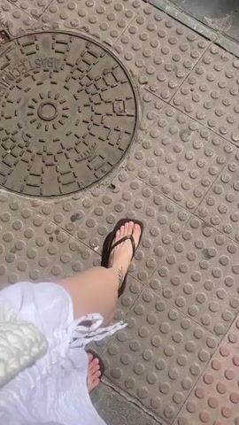 The video shows a first-person perspective of a person walking on a sidewalk. The person is wearing a white dress and black flip-flops. They are carrying a white textured bag in their left hand. The sidewalk is paved with large, dark gray square tiles, and there are circular textured tiles on the ground, possibly indicating a tactile paving surface for visually impaired pedestrians. A manhole cover with a patterned surface is also visible on the ground. The lighting suggests it is daytime with sunlight casting shadows. The camera angle is angled downwards, focusing on the ground and the person's legs and feet.