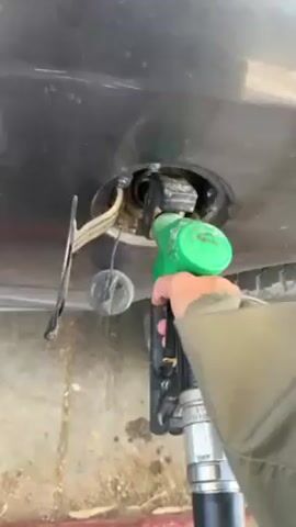 Here is a concise description of the video:

The video shows a first-person perspective of someone removing a green gas pump nozzle from a car's fuel tank, resulting in a spill of clear liquid. The car is dark in color. The ground beneath the car is stained. A spare tire can be seen next to the fuel tank.