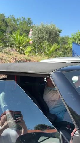 This is a close-up, vertical, interior shot of a black convertible car on a sunny day. The driver, a man wearing a light blue and white t-shirt and grey shorts, is visible in the driver's seat, looking to his right. The convertible top is partially open. A woman, holding a smartphone to take a selfie, is reflected in the car's windshield, wearing a white top. The background shows a lush green hillside with palm trees, foliage, and a road with traffic signs, suggesting a warm, outdoor environment.