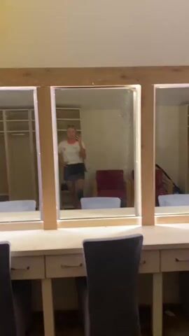 The video shows a young woman taking a mirror selfie in what appears to be a dressing room or backstage area. She is standing in front of a triple mirror with wooden framing and lights. She is wearing a white t-shirt with orange trim and a graphic logo, a black skirt, and sunglasses on top of her head. She is holding a smartphone in her right hand, taking the photo. A white bag is slung over her left shoulder. In the reflection, behind her, there are rows of lockers, red chairs, and other furniture, suggesting a communal space. The mirror surface is visibly dirty with water spots or dust. The overall impression is of a casual, behind-the-scenes moment.