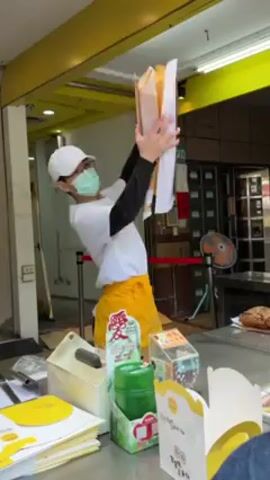 A person wearing a white cap, glasses, a blue face mask, a white shirt with dark sleeves, and a yellow apron is carrying a stack of five or six flat, square cardboard boxes. They are holding the boxes above their head, then lower them to a counter. The counter has various items on it, including boxes with yellow branding, a green container, a clipboard, and a pastry. The scene appears to be outdoors under a yellow awning with a building in the background.