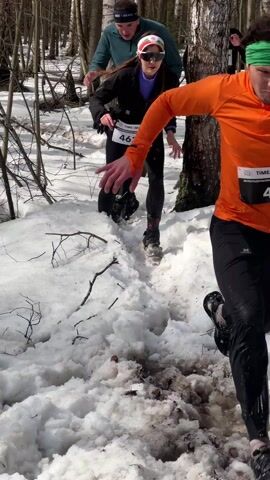 A group of runners participating in a race through a snowy forest is shown in the video. The camera angle varies from ground level to slightly higher, capturing the runners as they navigate a muddy and snowy path. The woman in the lead has a race number "462" visible on her chest, and is wearing sunglasses and a headband. The runners behind her are wearing similar race attire. They are running through a birch tree forest, which is covered with melting snow. The ground is a mix of mud, snow, and small sticks. The weather appears sunny, as trees are casting light shadows. An unseen person encourages the racers.