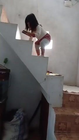 A young girl stands on a step of a staircase holding a tablet. She moves her arms and the tablet from side to side and then appears to be climbing or jumping down the stairs. The staircase has white steps and railing. A window with a cross-shaped light is visible behind the girl. In the foreground, a dark piece of furniture and some bags are visible. The floor tiles are brown.