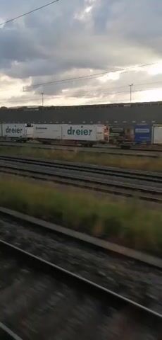 A train is moving past a railyard. The sky is cloudy and overcast with the sun attempting to break through. The railyard contains numerous train cars, many of which are yellow and appear to be carrying cargo. Some of the cars have company logos, including "dreier" and what appears to be a "P" logo. There are also some white cargo containers. Overhead power lines for the train are visible. The ground in the foreground is covered with grass and weeds, and several sets of train tracks are visible. The sound of the train moving is audible.