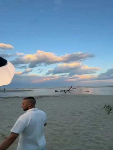 A man is standing on a sandy beach, posing for a photoshoot. He is wearing a white polo shirt and light blue shorts. A photographer in a green shirt and baseball cap is taking his picture with a professional camera.  A tall light stand with a white lantern-shaped light modifier is positioned between the subject and the camera. The background features a calm body of water with scattered driftwood and a sky filled with dramatic clouds at sunset.