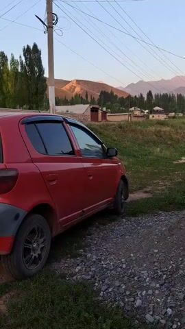 A red car is parked on a gravel and grassy area. The car is facing away from the camera, with its side visible. In the background, there are houses, trees, and mountains. The sky is clear and light.