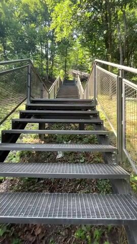 The video shows a close-up, upward-angled view of a long, metal staircase. The staircase is located outdoors, surrounded by lush green trees and foliage, suggesting a park or wooded area. The stairs have a grated metal surface and are flanked by metal railings. The sun is shining, casting shadows on the stairs and illuminating the leaves of the trees. The camera is positioned at the bottom of the staircase and appears to be moving slowly upwards, giving the impression of someone ascending the stairs. There are no people visible in the video. The overall scene is peaceful and natural.