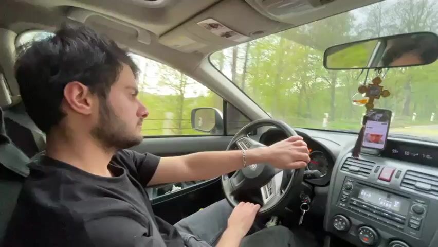 A young man with dark hair and a beard is driving a Subaru. He is wearing a black shirt and jeans. The car has a sunroof and a phone mount with a phone in it. A hand reaches in and runs fingers through the driver's hair.  The car is driving on a road lined with trees.  The audio includes music.