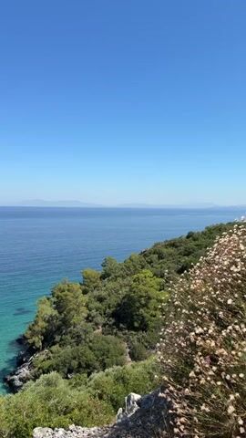 A vertical video shows a bright, clear blue sky over a calm, deep blue sea. The water near the coast is a vibrant turquoise, revealing rocky shores and a lush green hillside covered in trees and bushes. Distant islands are visible on the horizon. The camera pans slightly to the right. There are no people or man-made objects visible.