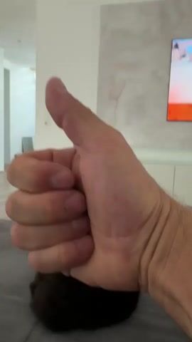 The video shows a close-up of a person's right hand giving a thumbs-up gesture. The hand is in the foreground, with the thumb extended upwards and the fingers curled into a fist. The background is blurred, but appears to be an indoor setting with a couch and a television screen displaying a colorful image. A dark, furry object, possibly a pet, is partially visible beneath the hand on the couch. The lighting is bright, and the overall impression is that of a casual, informal moment.