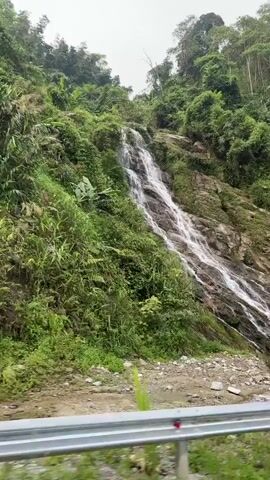 This video captures a scenic view from a moving vehicle traversing a winding road in a lush, mountainous region, likely Tân Quang Commune, Vietnam.

The main subject is a beautiful waterfall cascading down a rocky, green-covered slope on the right side of the frame. The water flows over several tiers of rocks, creating a dynamic visual. The entire hillside is densely vegetated with various shades of green foliage, including trees, bushes, and smaller plants, indicating a vibrant, possibly tropical or subtropical, ecosystem.

In the foreground, a paved road with yellow markings is visible, curving gently to the left. A metal guardrail runs alongside the road, separating it from a drainage ditch and the natural slope. Part of a vehicle's side, possibly a side mirror or frame, can be seen in the lower right corner, indicating the perspective is from inside a car or motorcycle.

The sky appears overcast, suggesting a cloudy day, but there is sufficient light to illuminate the landscape clearly. The overall impression is one of natural beauty and a tranquil journey through a verdant environment. No people or visible text are present in the video.