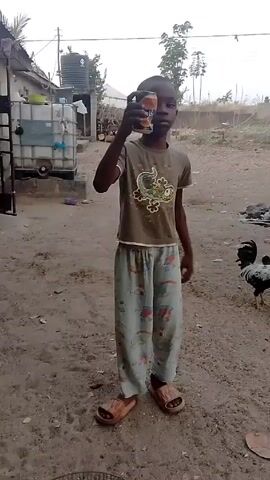 A young boy stands in a dirt yard, holding a can. He is wearing a brown t-shirt with a cartoon on it, pajama pants, and sandals. Behind him, several chickens peck at the ground. The boy drinks from the can.  There is a water tank and various objects scattered around the yard.