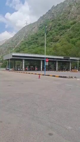 A low angle shot shows a toll plaza with a green mountain looming behind it. The plaza is a rectangular structure with a flat roof, supported by numerous columns, each marked with a stop sign. A few vehicles, including an orange truck and a motorcycle, are seen under the plaza's roof, possibly paying tolls. The road in front of the plaza is wide and grey, marked with white lines and a few orange traffic cones. The mountain is covered in green vegetation, with a rocky peak visible in the background. An electric transmission tower is partially visible on the mountain. The sky above is partly cloudy, suggesting a bright, sunny day.