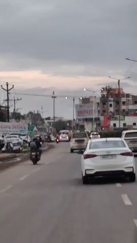 The video shows a view from a vehicle traveling on a paved road in what appears to be a city in India. The sky is overcast. On the left side of the road, there are shops and parked vehicles, including motorcycles and cars. There are also streetlights and utility poles. A green directional signpost indicates "Paria Phata" and "Dhanori". Several people are visible on the sidewalk. Ahead in the lane are various vehicles, including motorcycles, cars, and a bus. A white sedan is prominently in the foreground on the right side of the frame. The overall impression is of a busy urban street scene.