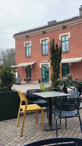 The camera pans up to show a brick building with a sign reading "NALEJ SE" above the door. The building has green windows and a green door. There are tables and chairs outside the building. A bicycle is parked nearby. The sky is overcast.