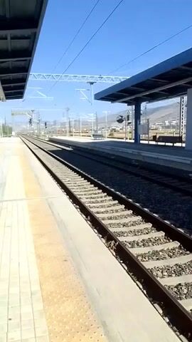 The video shows a train station with clear blue skies and a bright sun. The camera pans from left to right, focusing on train tracks and the platform. On the platform, there are blue-roofed shelters, support pillars, a bench, and what appears to be an elevator. In the distance, mountains can be seen, along with more train tracks and some buildings. The train tracks are well-maintained with ballast stones. The video is taken from a stationary perspective, possibly from another train or a platform. There are no people visible in the video. The overall scene depicts a modern and functional train station in an outdoor environment.