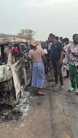 The video shows a burnt-out van on the side of a road. A group of people are standing around the van, looking at it. One woman is reaching into the van. The road is paved and there is vegetation on either side of the road. The weather appears to be hazy.