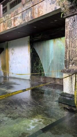 The camera pans across a large, deteriorated, and possibly abandoned building. The architecture has a traditional Southeast Asian style, with columns and multi-tiered balconies. The building shows signs of significant water damage, with mold and staining visible on the walls and columns. The floor is wet, reflecting the light.  Yellow caution tape is stretched across the foreground, likely indicating that the area is off-limits or dangerous. The scene suggests neglect and disuse.