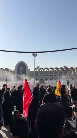 The video shows a large crowd of people gathered outdoors, under a bright blue sky. Many people are wearing dark clothing. Several flags are visible, including a prominent red flag with writing on it and an Iranian flag. In the background, there is a large building with arched windows and a portrait of a man in traditional attire displayed prominently on its facade. Water is being sprayed in the air in the background, creating a mist. The crowd appears to be moving, with some individuals holding flags and others observing or participating in an event. The overall atmosphere seems to be one of a public gathering or procession.