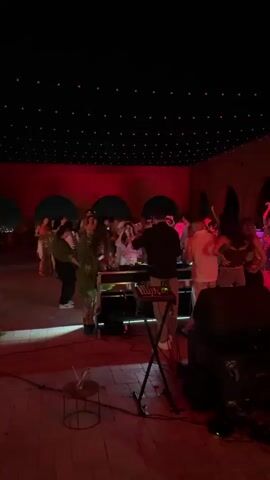 The video shows an outdoor rooftop party at night. A DJ is set up in the center of the frame, surrounded by people dancing. The scene is lit by red lights and string lights overhead, creating a vibrant atmosphere. Some people are dressed casually, while others are more dressed up for the occasion. The architecture of the building suggests a Mediterranean or Spanish influence, with arches visible in the background. The overall impression is one of a lively and energetic gathering.