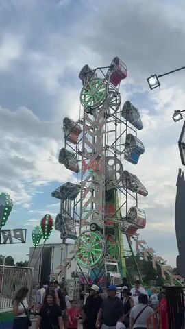 The video shows a tall amusement park ride at a fair with multiple gondolas. The ride is illuminated with colorful LED lights that change pattern and color throughout the clip. People are gathered at the base of the ride, some standing in line and others watching. The sky is cloudy and appears to be late afternoon or early evening. There are also other fair attractions visible in the background, including what appear to be hot air balloons and other rides. The overall atmosphere is lively and festive.