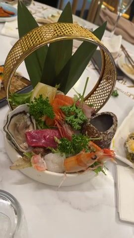 A white table, with a marble pattern, holds a serving of oysters, raw fish, and shrimp in a white bowl with a gold handle. There are green sprigs of herbs as well as a lemon wedge. Next to it is a small dark bowl with a darker sauce. Chopsticks are seen near other bowls and plates. A tray of sushi sits to the left. Wine bottles in ice sit in front of a woman at the table, who is facing away from the camera. A black wall with a white logo is behind the table. Several empty glasses are on the table.