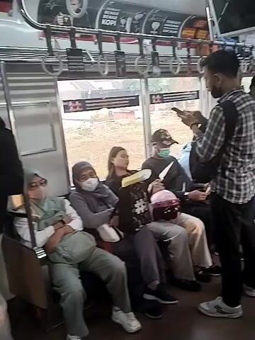 The video shows the interior of a train with several passengers. In the foreground, a man in a checkered shirt is standing and looking at his phone. He is holding a bag over his shoulder. To his left, several people are sitting on a bench. One woman wearing a hijab and glasses is looking down with her arms crossed. Next to her, another woman, also wearing a hijab, is looking down and appears to be sleeping. A woman with long hair is sitting beside them, looking forward. A man wearing a cap and a mask is seated next to her. In the background, more passengers are visible, some sitting and some standing. The train appears to be moving, as indicated by the scenery visible through the windows. There are handrails and overhead compartments typical of public transportation.