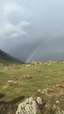 A sweeping vertical shot showcases a vibrant rainbow over a mountainous landscape. The foreground features a grassy field dotted with clusters of rocks, some covered in lichen. In the distance, a small settlement nestles within the valley. The rainbow arcs dramatically against a backdrop of gray, overcast skies, culminating near a snow-capped mountain range. The camera pans slightly, revealing additional terrain including rugged rocky slopes covered with brown grass.