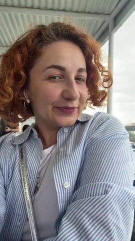 A woman with red curly hair is seated on a blue plastic chair on what appears to be a boat or ferry. She is wearing a white shirt under a blue and white striped button-up shirt, with a silver crossbody bag. She is smiling and looking at the camera.  Behind her, through the railing, is a view of a city with buildings of various sizes, some trees, and a waterfront area with yellow markings. The sky is overcast.  The camera pans slightly to the right to show more of the scenery.