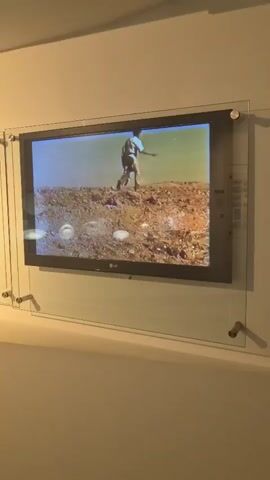 A video plays on a flat-screen television mounted on a wall. The video shows a person from the waist up, wearing a light-colored shirt and dark pants, walking across a dry, rocky terrain under a bright sky. The person is walking away from the camera. The terrain is covered in small rocks and dirt. The video is framed by a clear acrylic panel held to the wall by four metal screws. To the left of the television, there is a display of what appear to be postage stamps mounted on a lighter colored panel, also enclosed in acrylic. The wall is painted a neutral color.
