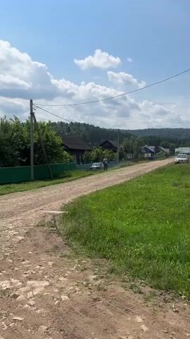 A dirt road winds through a grassy landscape under a bright blue sky with scattered clouds. On the left, a green fence borders a line of trees. A utility pole with wires stands near the fence. In the middle ground, a few wooden houses are visible along the road, surrounded by vegetation. A person on a bicycle rides down the road, moving away from the camera. The overall scene depicts a rural environment on a sunny day.