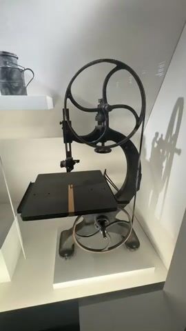 The video shows a vintage sewing machine on display. The machine is dark-colored with large wheels and a belt-driven mechanism. The sewing platform is flat and black, with a strip of orange tape on it. To the left of the sewing machine, a portion of a metal jug is visible on a white shelf. The machine is displayed on a white platform, possibly in a museum or exhibition setting, with a white background and subtle lighting. The video is filmed from a medium distance, capturing the entire sewing machine and its immediate surroundings.