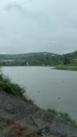 A video captures a view through a rain-streaked window, looking out at a body of water bordered by green hills under a cloudy sky. A dark pole with a yellow warning sign is visible in the foreground on the left. The camera appears to be moving slowly, indicated by the shifting perspective of the landscape and the movement of raindrops on the glass. The scene is predominantly green and grey, suggesting overcast and wet weather conditions. No people or specific actions are discernible.