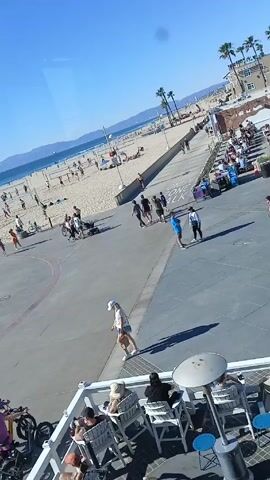 Here's a concise description of the video:

The video shows a sunny beach scene from an elevated perspective, likely through a window. The beach is crowded with people playing volleyball and walking. A paved area is adjacent to the beach, with people strolling and biking. Some are seated at an outdoor restaurant or patio area visible in the foreground. Palm trees dot the landscape. The ocean is visible in the background, with distant mountains on the horizon.  The American flag is visible on the left side of the beach area.