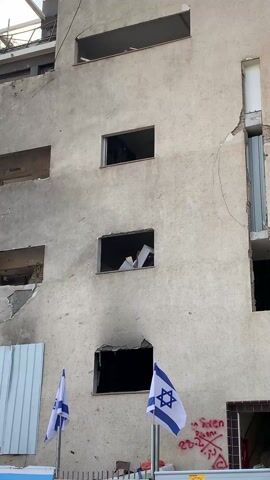 The video shows a damaged building with several holes in the exterior walls. A few Israeli flags are visible in front of the building. There is a metal fence and some construction signs present. The building appears to be under construction or demolition. The sky is overcast, and sunlight is filtering through the clouds. A building nearby appears to be similarly damaged.