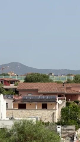 The video shows a daytime exterior scene with a clear, light blue sky. In the foreground, there are several buildings with terracotta tiled roofs, and some trees and foliage. Some of the buildings have solar panels installed on their roofs. In the middle ground, there are more buildings and dense green trees. In the background, there are hills and mountains, and a construction crane is visible on the right side of the frame. A road is visible on the right side with two cars driving on it. The overall impression is a residential area with some ongoing construction.
