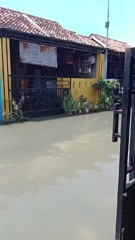 The video shows a flooded residential street. The water level reaches up to the base of the houses, submerging the roads and sidewalks. The houses are closely spaced, with a mix of yellow and blue exteriors. There are a few potted plants visible near the entrance of one house. A man wearing a red shirt sits in a chair in the open doorway of a blue house. Another person, also wearing a red shirt, stands near the gate of a yellow house. The sky is visible and appears bright. There is a black metal gate in the foreground, slightly ajar, obscuring a portion of the view.