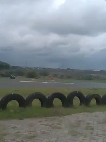 The video shows a shot of a racetrack with two motorcycles racing on it. There are tires lining the side of the track in the foreground. The sky is overcast with dark, threatening clouds. The motorcycles are small in the frame and appear to be moving at high speed. The background shows a flat, grassy landscape with sparse vegetation and what looks like a town in the distance.