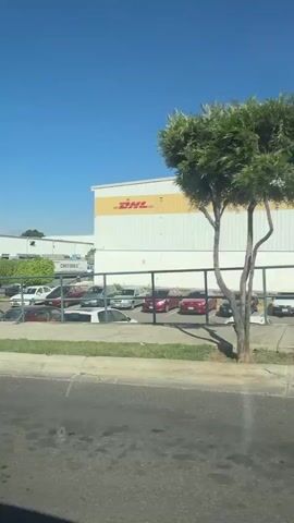 The video provides a drive-by perspective of an industrial area in Sanctorum, Mexico, bathed in bright sunshine under a clear blue sky.

The scene begins with a view of a large white building, clearly marked with the red and yellow DHL logo, with a parking lot in front of it containing numerous cars of various makes and colors. As the camera pans to the left, more industrial buildings become visible in the background. The foreground features a green metal fence separating the road from more parked cars and neatly trimmed green trees. A red circular sign with a diagonal line over the letter 'E' (indicating "No Parking") is briefly seen.

The camera continues to move, showing another long, white industrial building. In front of this building, several vehicles are parked, including a black pickup truck, a silver sedan, and a large white semi-trailer truck further down the road. The video concludes with a shot of a man walking on a sidewalk with a backpack, passing by another "No Parking" sign. He is walking alongside a grassy slope and various trees, including some that resemble palm trees, before the camera moves past him. The overall setting depicts a busy industrial or commercial district.